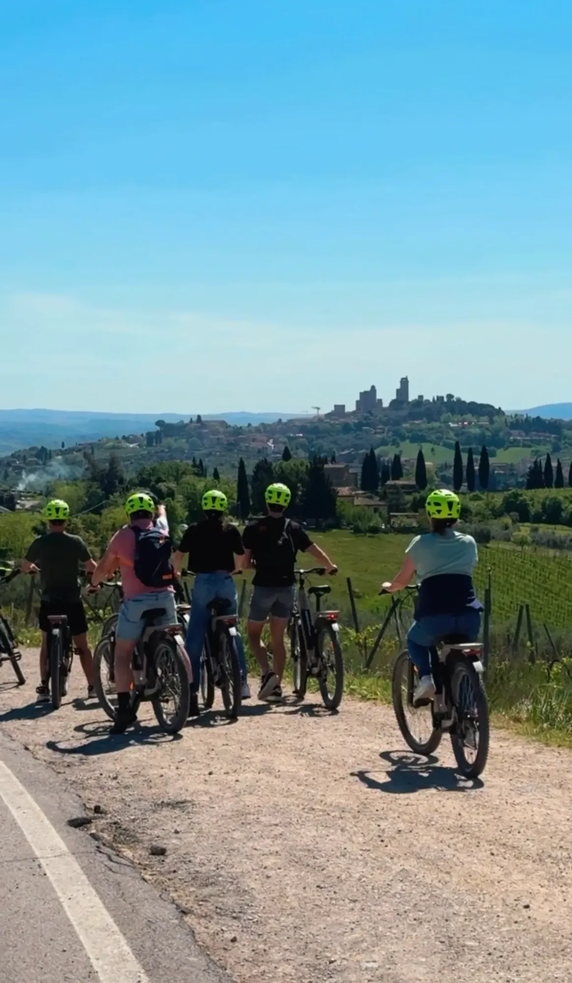 A happy e-bike group, helmets on, waving on a white gravel road through the Tuscan vineyards