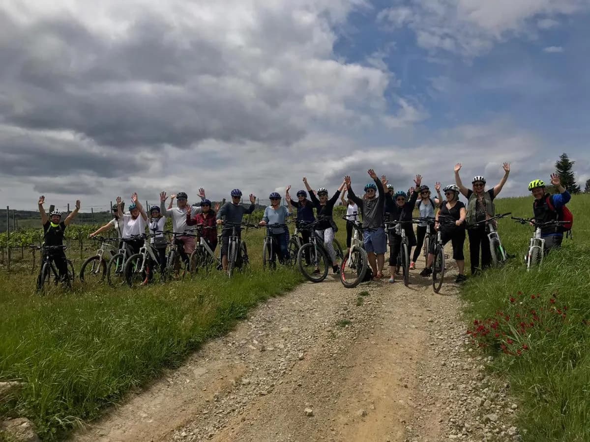 A happy e-bike group, helmets on, waving on a white gravel road through the Tuscan vineyards