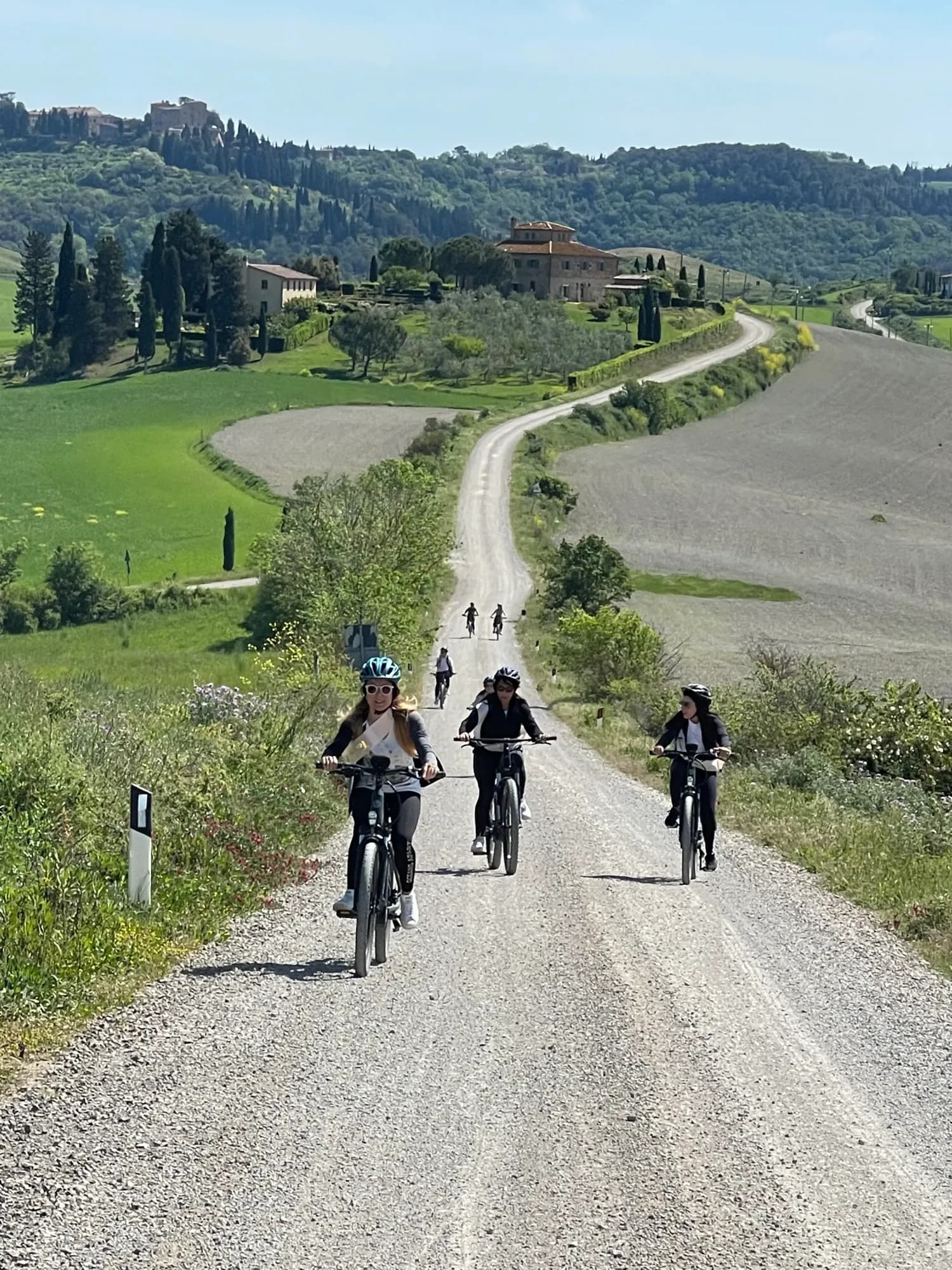 Three guests e-biking a white gravel road below a Tuscan farmhouse and cypress trees